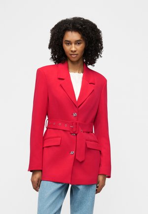 Femme aux cheveux bouclés portant un blazer rouge vif ceinturé sur un haut blanc et un jean bleu clair, debout devant un fond uni.