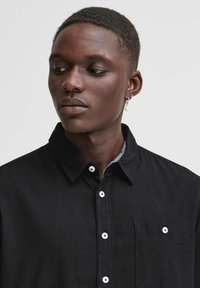 Svart denimskjorte med knappekrage, lommedetalj og hvite knapper. Modellen har kort hår og en perleøredob.