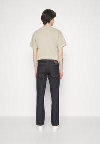 Beige katoenen t-shirt, korte mouwen, gecombineerd met donkerblauwe denim jeans. Witte sneakers zichtbaar aan de onderkant. Effen achtergrond.