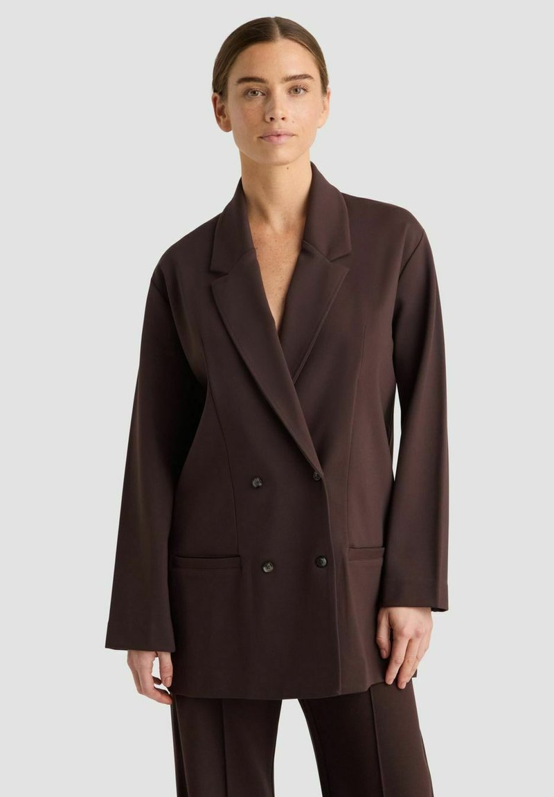 Eine Frau trägt ein dunkelbraunes, doppelt geknöpftes Blazer mit passender Hose und steht vor einem schlichten hellgrauen Hintergrund.