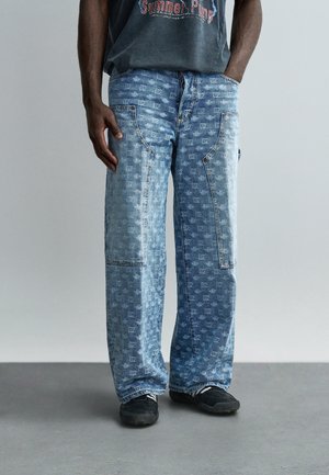 Hombre con pantalones vaqueros de mezclilla azul claro con bolsillos parche y un sutil patrón cuadrado, combinados con zapatillas negras y una camiseta oscura con gráfico.