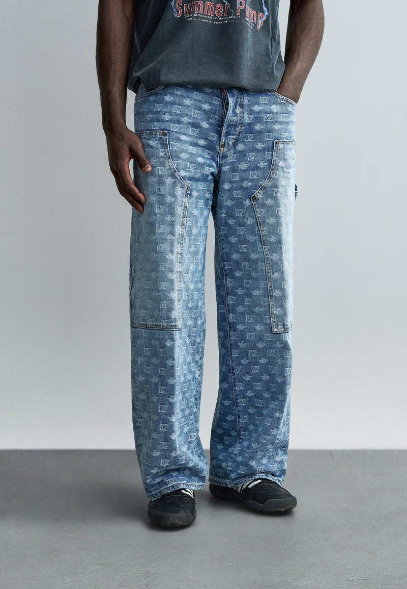 Άνδρας που φοράει ανοιχτό μπλε τζιν παντελόνια με τσέπες με πατς και λεπτό τετράγωνο μοτίβο, συνδυασμένα με μαύρα αθλητικά παπούτσια και σκούρο μπλουζάκι με στάμπα.