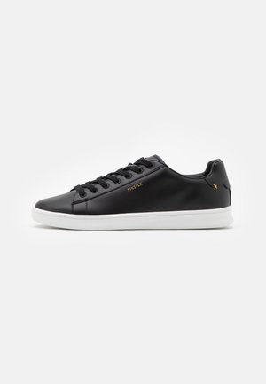 Baskets basses noires avec semelle blanche, lacets noirs, logo doré "SIKSILK" sur le côté et petit emblème doré près du talon.