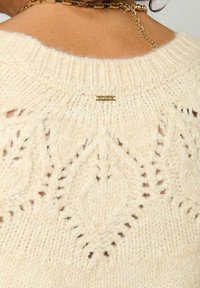 Pull en tricot de couleur crème avec un motif en losanges, un fil texturé et une petite étiquette dorée près de l'encolure.