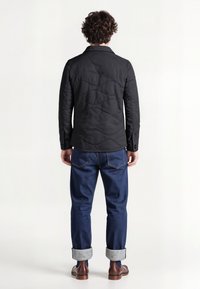 Sort quiltet jakke med krave og knapmanchetter, parret med optrillede blå denim jeans og mørkebrune sko, set fra ryggen.