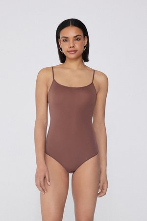 Frau mit glattem schwarzen Haar, die einen braunen ärmellosen Bodysuit trägt und vor einem schlichten hellgrauen Hintergrund steht.