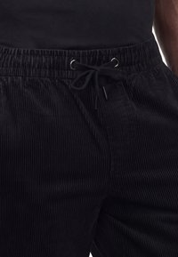 Man wearing black corduroy pants with a drawstring waist, viewed from the waist to mid-thigh, paired with a black shirt.