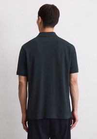 Donkergroene polo, korte mouwen, gladde stof, rechte zoom en subtiele kraagdetails. Minimalistisch ontwerp zonder zichtbare logo's of patronen.