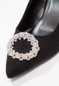 Escarpin noir à bout pointu avec une grande boucle ronde en strass argenté sur le devant.
