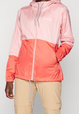 Persona vistiendo una chaqueta con capucha Columbia bicolor en tonos rosa y coral, con cordones, cremallera frontal, y pantalones beige para exteriores.