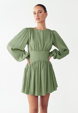 Femme aux cheveux plaqués en arrière, portant des boucles d'oreilles dorées et une robe vert clair à manches longues bouffantes, mains sur les hanches, fond neutre.