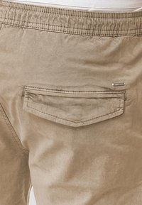 Shorts couleur beige avec une ceinture élastique, dotés d'une poche arrière rectangulaire et de détails de couture discrets. Tissu léger avec une texture lisse.