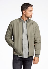 Chaqueta bomber acolchada en verde oliva con puños acanalados, cierre de cremallera en la parte delantera y bolsillos laterales, superpuesta sobre una camisa de botones de color gris claro.