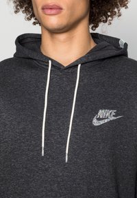 Mörkgrå hoodie i mjukt material. Har en vit dragning och en glittrande Nike-logotyp på bröstet. Enkel, avslappnad design.