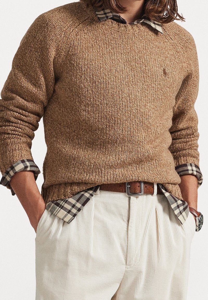 Personne portant un pull en laine marron sur une chemise à carreaux avec les manches retroussées, un pantalon en velours côtelé blanc, une ceinture en cuir marron et une montre-bracelet, les mains dans les poches.