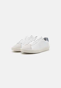 Une paire de baskets blanches basses avec des embouts en daim gris clair et des accentuations bleues sur les languettes de talon, des semelles en caoutchouc beige et des lacets blancs.