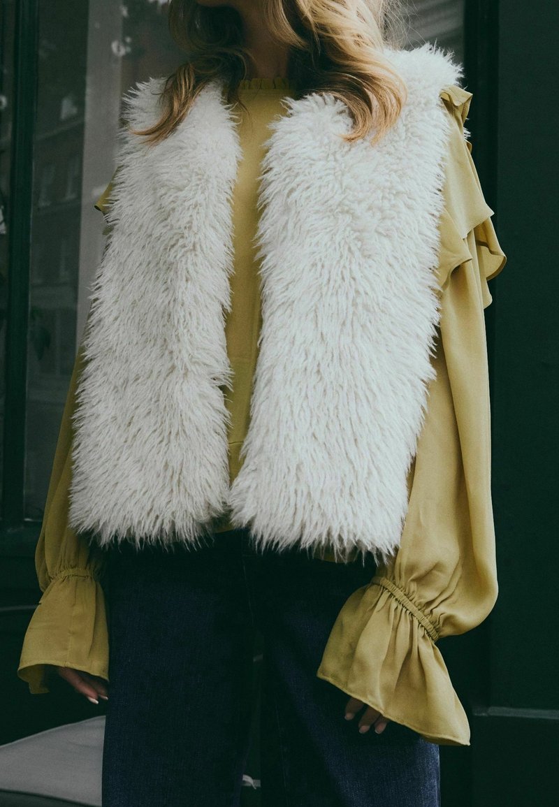 Femme portant un gilet blanc duveteux par-dessus un chemisier jaune moutarde à manches longues avec des poignets à volants et un pantalon foncé, debout près d'une fenêtre.