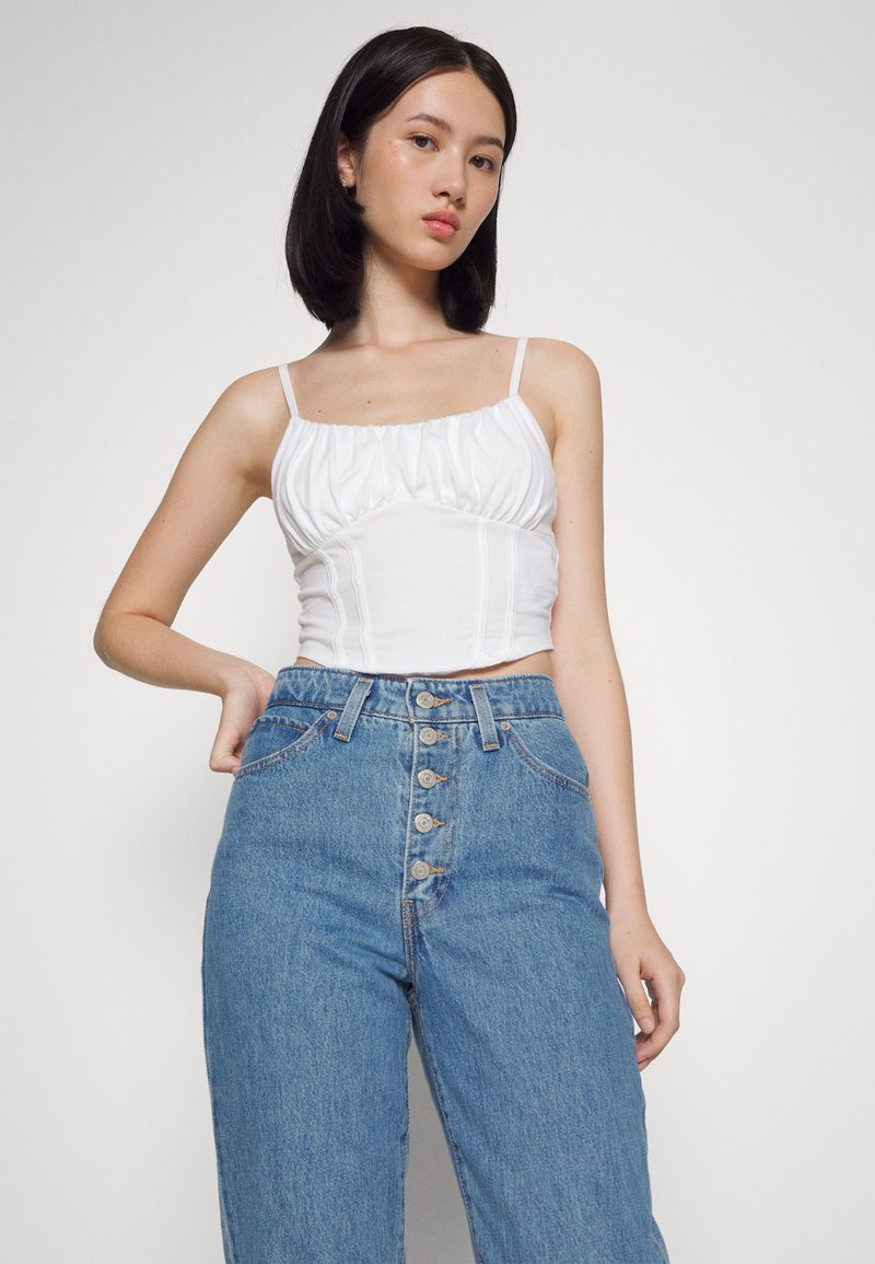 Top blanco corto con cuello rizado, combinado con jeans de mezclilla azul de cintura alta que cuentan con un frente con botones y bolsillos.