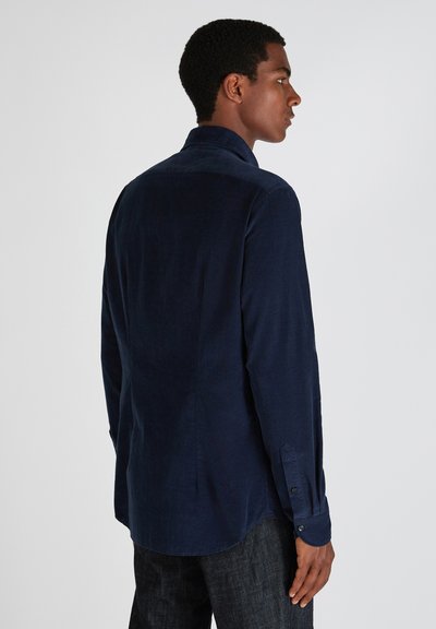 Man in een donkerblauwe lange mouw shirt en donkere jeans, in profil staan tegen een effen lichte achtergrond.