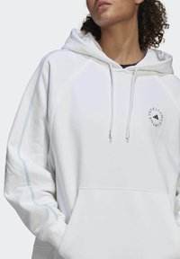 Person med en vit hoodie med ljusblå sömmar på ärmarna och en rund svart logotyp på bröstet, händerna i framfickan.