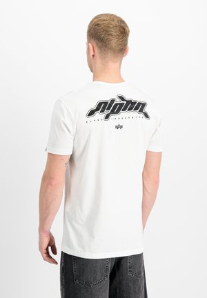 Man with short blond hair wears white t-shirt with large black "Alpha Industries" logo on upper back, paired with dark gray jeans.