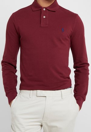 Hombre vestido con una camisa polo de manga larga color burdeos con botones blancos y pantalones color crema, con las manos en los bolsillos.