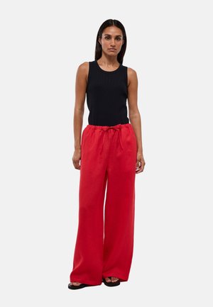 Femme portant un haut noir sans manches, un pantalon large rouge avec une taille à cordon et des sandales noires ouvertes à l'avant, debout devant un fond uni.