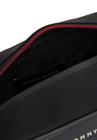 Black fabric pouch with red zipper, featuring a textured surface. Inside, a label reads "TOMMY HILFIGER," with a zip pocket visible.