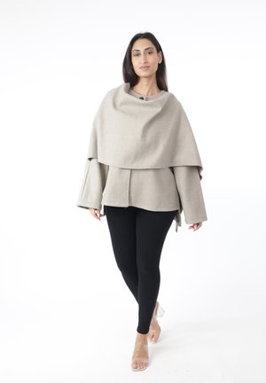 Woman wearing a light beige layered cape coat, black leggings, and clear high-heeled sandals, standing on a white background.