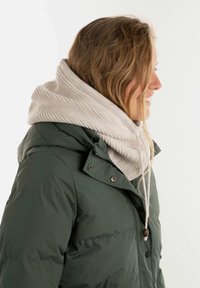 Groene pufferjack met een hoge kraag, gecombineerd met een lichtbeige geribbelde brei-sjaal. De sjaal heeft een koord met een houten stopper.