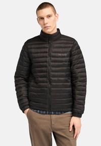 Giacca imbottita nera con colletto alto, presenta un design a righe orizzontali, chiusura a zip e logo Timberland sul petto. Vestibilità casual.