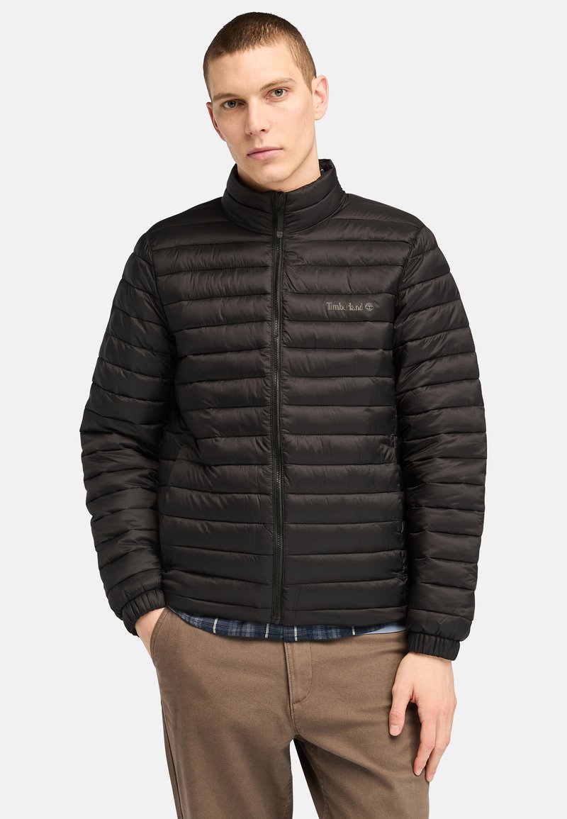 Giacca imbottita nera con colletto alto, presenta un design a righe orizzontali, chiusura a zip e logo Timberland sul petto. Vestibilità casual.