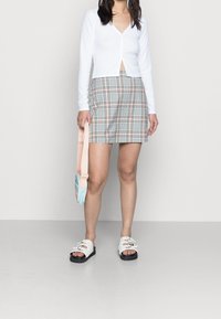 Bílý žebrovaný top s dlouhým rukávem, kostkovaná mini sukně v růžové, modré a béžové barvě, plastová crossbody taška a bílé sandály s černými podrážkami.