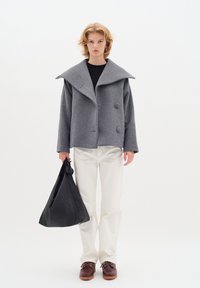 Graue Wollmischungjacke mit übergroßem Kragen und zwei Knöpfen, kombiniert mit weißen Hosen und einer schwarzen Lederhandtasche. Model trägt braune Schuhe.