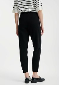 Schwarze, schmal zulaufende Hose mit einem elastischen Bund, weicher Textur und dezentem Glanz, kombiniert mit schwarzen Slippern, von hinten gesehen.