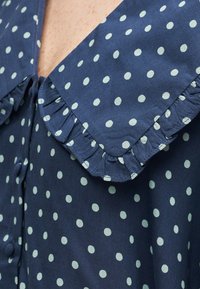 Blouse bleu marine à pois blancs avec un grand col carré à bords froncés et des boutons recouverts de tissu.