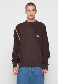 Pull en tricot marron foncé avec un motif diagonal, une bande d'accent orange et un petit logo sur la poitrine. Coupe décontractée.