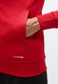Κόκκινο φούτερ με υφή, που διαθέτει πλαϊνή τσέπη, ριμπ μανσέτες και ορατό λογότυπο. Σχεδιασμένο για αναπνοή και άνεση.