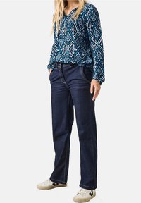 Blaue gemusterte Bluse mit langen Ärmeln, kombiniert mit dunkelblauen Jeans und weißen Sneakers. Die Bluse besitzt ein Diamantdesign in unterschiedlichen Farbtönen.