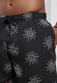 Bañador negro con motivos de sol bordados en blanco, cintura con cordón ajustable y bolsillos laterales. Tejido ligero con textura suave.