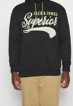 Czarna bluza z kapturem z zielonymi sznurkami i dużym napisem z przodu: "Jack & Jones Superior Est. 90 Intl", noszona z beżowymi spodniami.