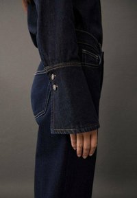Personne portant une veste en jean bleu foncé et un jean assorti, vue de profil avec une main détendue sur le côté devant un fond gris uni.