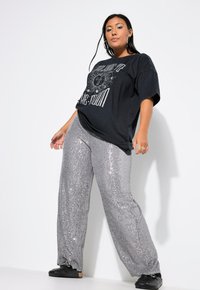 T-shirt nera over size con stampa grafica, abbinata a pantaloni di paillettes argento lucido. Modella che indossa scarpe nere, in piedi su uno sfondo bianco.