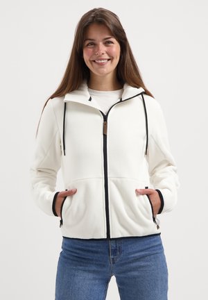 Eine junge Frau lächelt, trägt einen weißen Zip-Hoodie mit schwarzem Besatz und blaue Jeans, die Hände in den Taschen des Hoodies, vor einem schlichten Hintergrund.