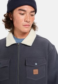 Fleece-takki tummansinisellä värillä, jossa on kerma väri kauluksessa, vetoketjusulku, kaksi rintataskua ja merkkikohtainen nahkapatchi. Pehmeä tekstuuri, rento muotoilu.