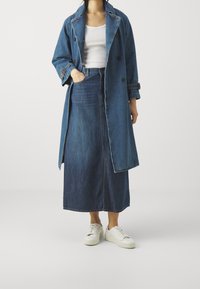 Trench coat in denim blu scuro con una vestibilità rilassata, sovrapposto a un top bianco e una gonna in denim blu coordinata, abbinato a sneakers bianche.