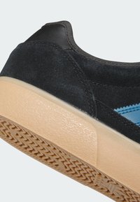 Čierna semišová teniska s gumovou podrážkou. Má modrý akcent a textúrovaný povrch. Zadná časť zobrazuje kožený oporný pás a vzorovanú vonkajšiu podrážku.