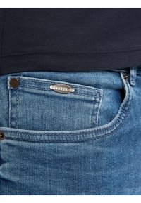 Jeans de mezclilla azul claro con un acento de logo plateado en el bolsillo trasero y una textura de lavado medio. Presenta un botón negro en la cinturilla.