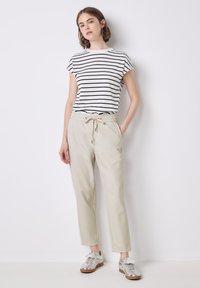 Vrouw staat tegen een witte muur, gekleed in een kortebeurs witte shirt met zwarte strepen, beige broek met koord en zilveren sneakers.