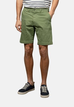 Hombre con pantalones cortos de color verde oliva hasta la rodilla y zapatillas negras bajas con cordones blancos, de pie con una mano en el bolsillo.